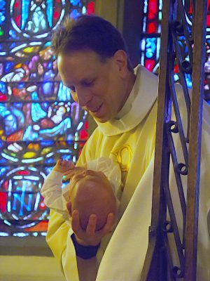 Adam Trambley holding an infant at baptismal font in front of stained glass window